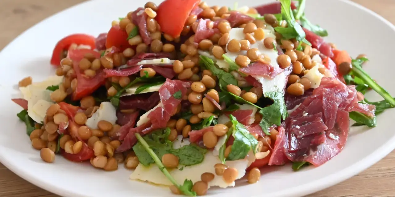 Insalata di lenticchie con pomodorini, scaglie di parmigiano e prosciutto crudo, un piatto fresco e gustoso.