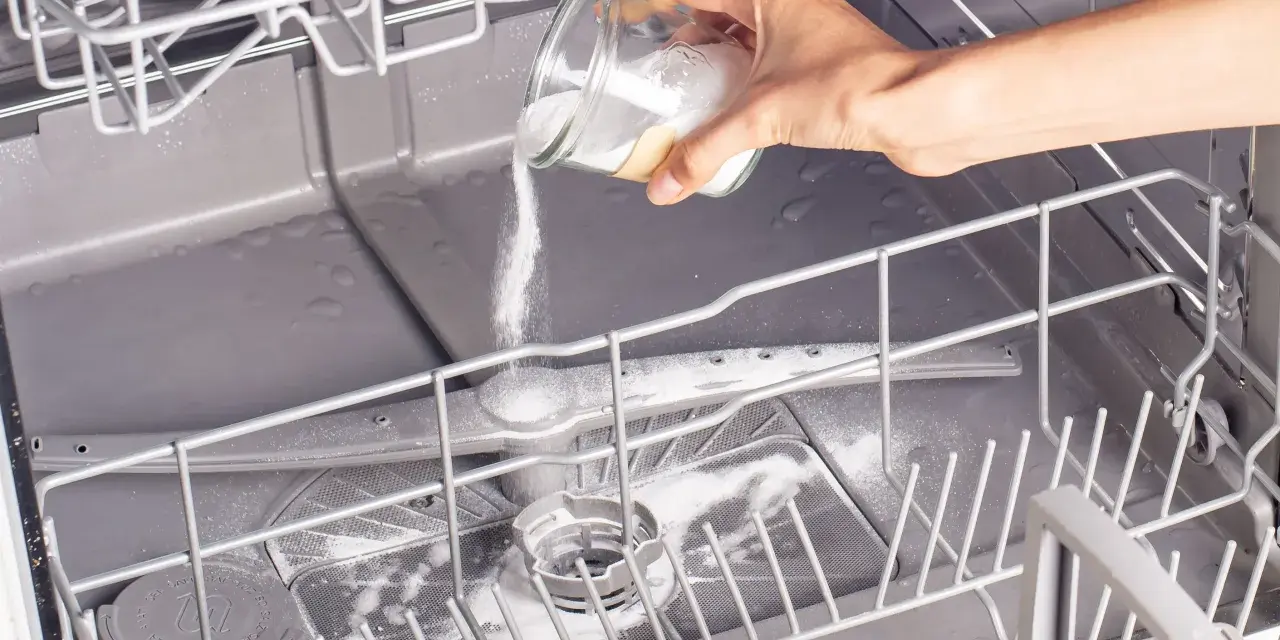 A hand pours white powder from a jar into an open dishwasher, demonstrating how to clean a dishwasher naturally.
