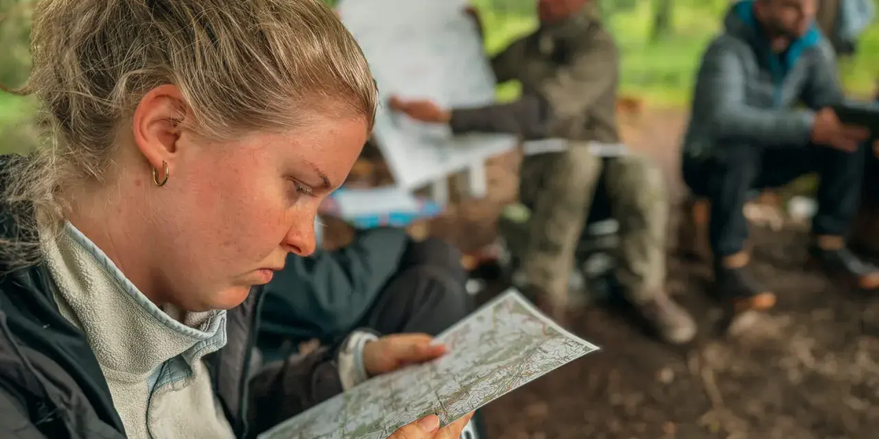 Frau studiert Karte, während Männer im Hintergrund bei einer Ausbildung im Wald sitzen. Solche intensiven, kurzen **ausbildungen die nur 1 jahr dauern** sind ideal für schnelle Karriereschritte.