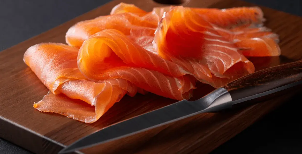 Finas lonchas de salmón ahumado, listas para disfrutar, reposan sobre una tabla de madera junto a un cuchillo.