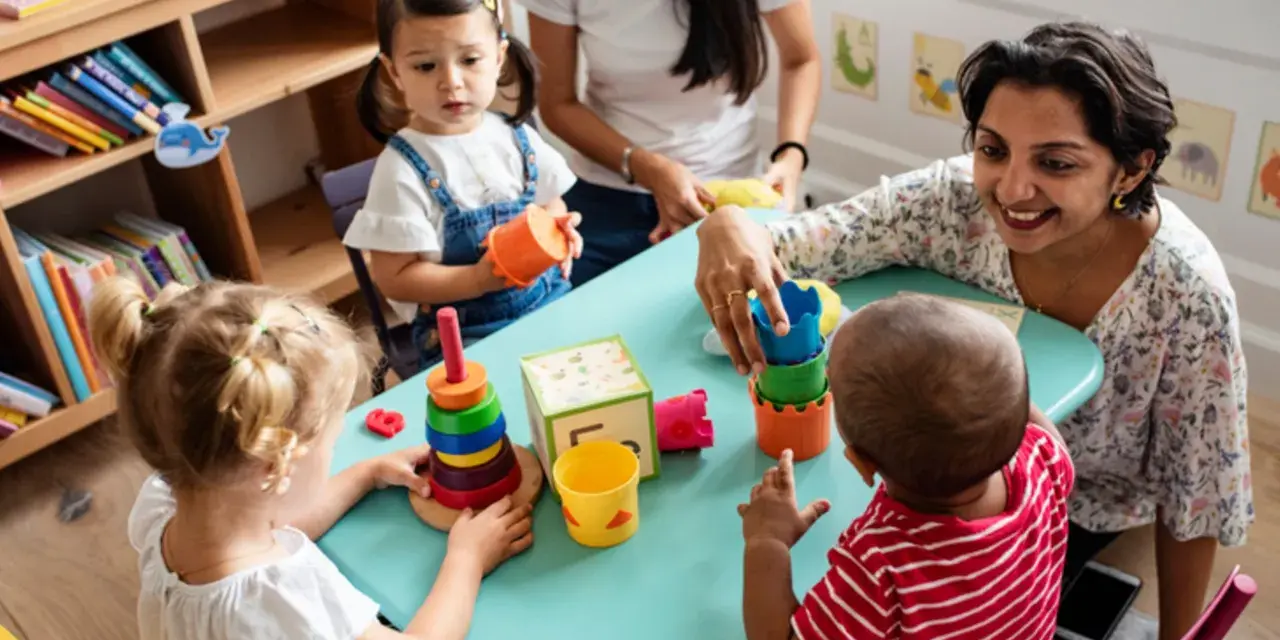 Zwei Frauen und drei Kleinkinder spielen mit bunten Spielsachen. Eine Frau hilft einem Kind, Türme zu stapeln, was eine tolle Methode ist, um Kinder zweisprachig zu erziehen.