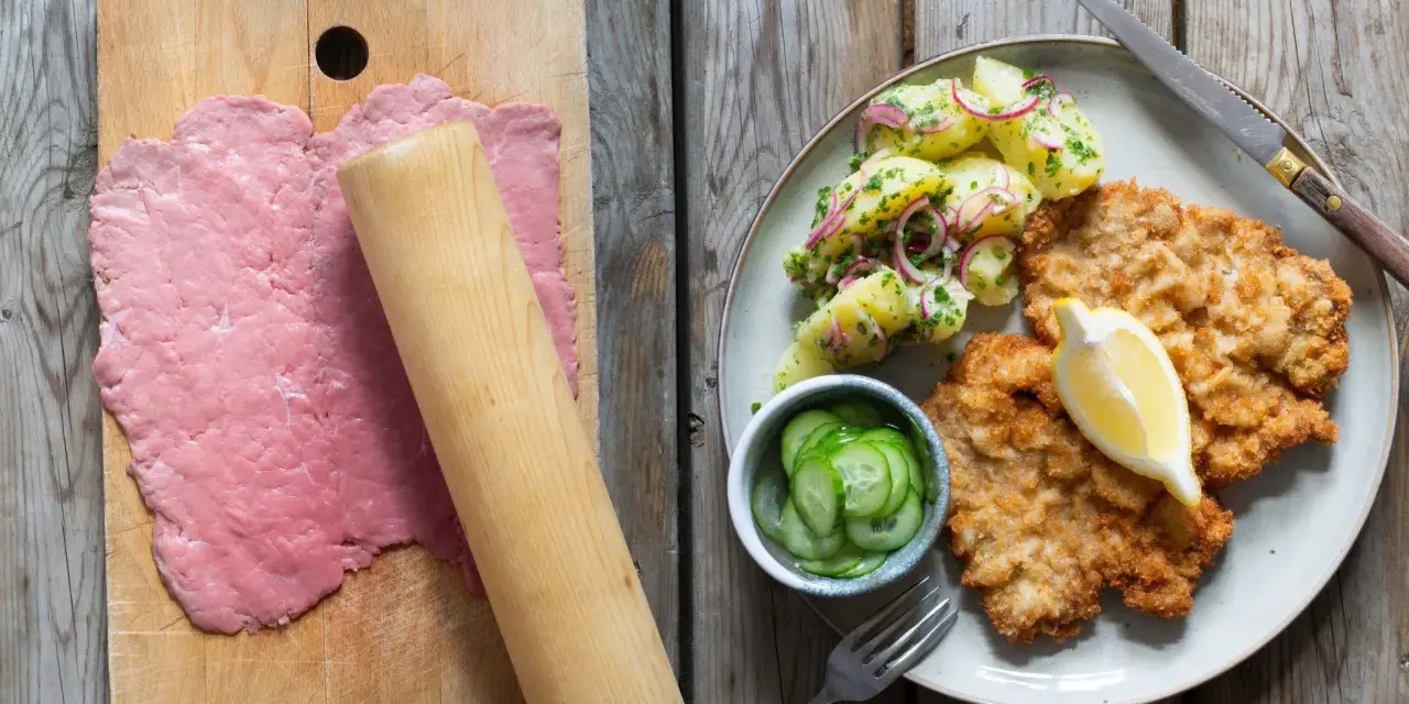 Förberedelser för schnitzel recept: tunt bankat kött på skärbräda, bredvid en tallrik med schnitzel, potatissallad och gurka.