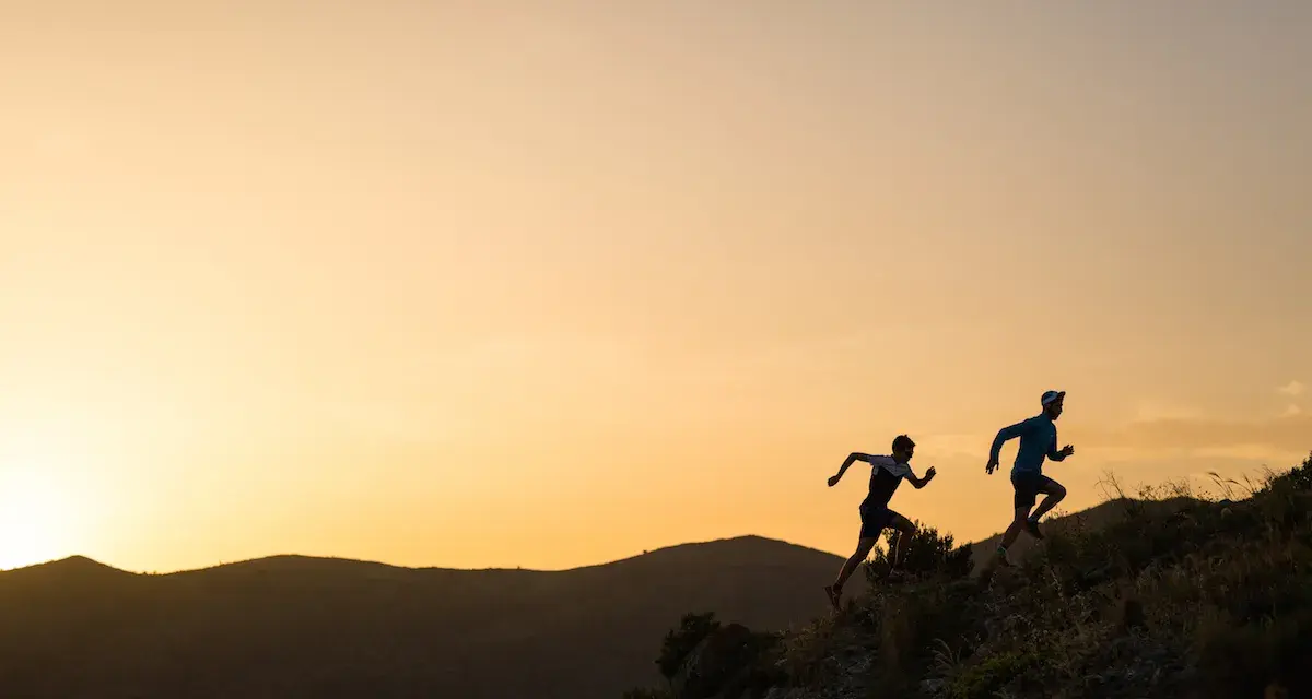 Dos corredores se enfrentan a una exigente carrera de senderos al amanecer, sus siluetas recortadas contra un cielo anaranjado.