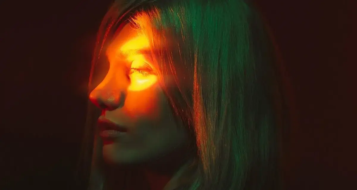 Profilo di donna con luce arancione sull'occhio, evocando un'emicrania oculare. Capelli illuminati da luci verdi e rosse.