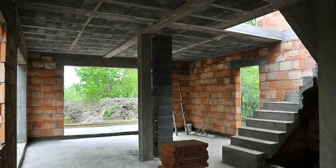 Wnętrze budowy z widocznym stropem gęstożebrowym, schodami, ceglanymi ścianami i otwartymi drzwiami na zewnątrz.