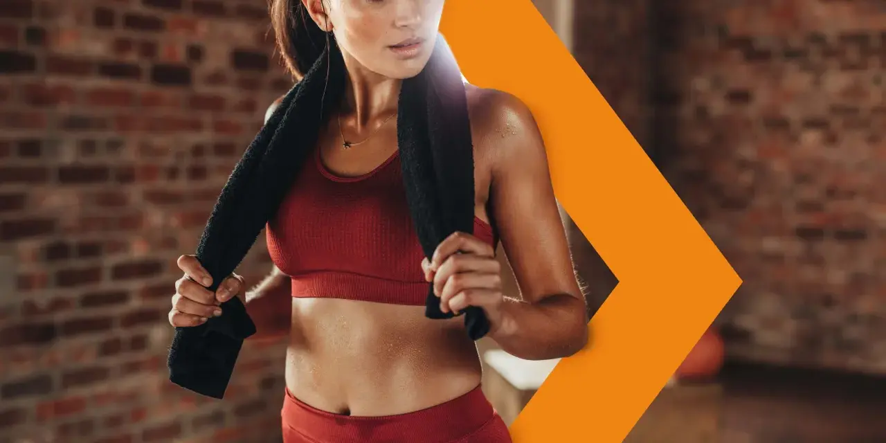 Donna sudata in abbigliamento sportivo rosso, con un asciugamano nero, si riposa dopo l'allenamento. Con il caldo i battiti aumentano, ma lei è determinata.