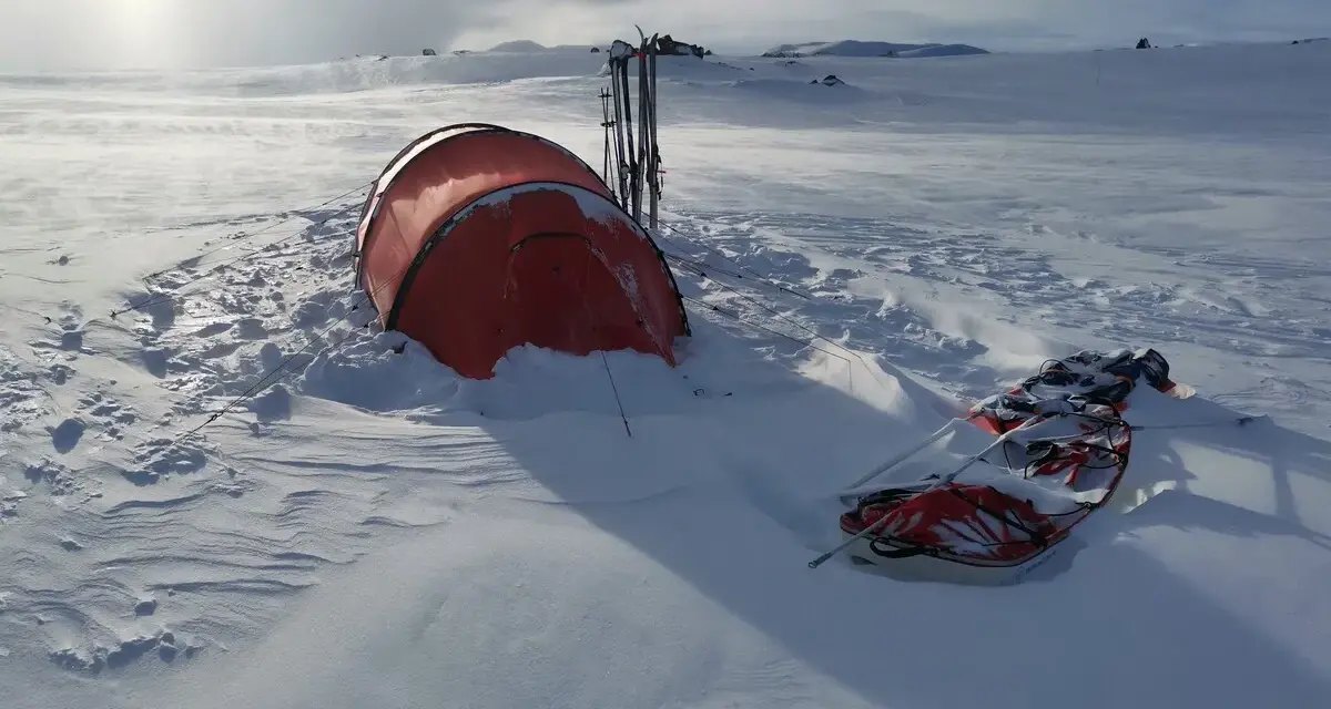 Ein rotes Wintercamping Zelt steht im Schnee, daneben Skier und ein Schlitten. Die Landschaft ist weit und verschneit.
