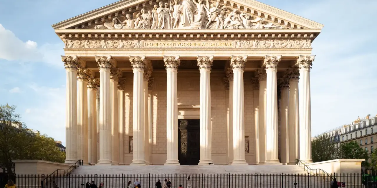 La imponente iglesia de la Madeleine en París, con sus columnas neoclásicas y esculturas en el frontón, se alza bajo un cielo azul.