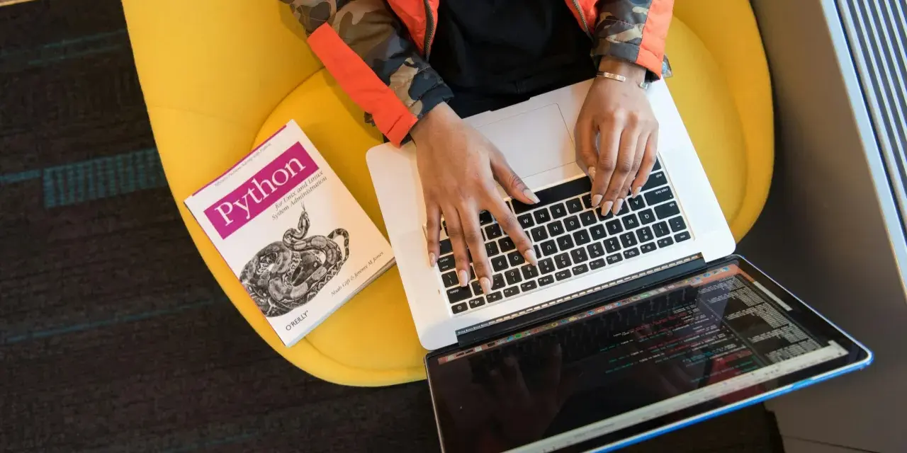 Osoba w pomarańczowej kurtce pisze na laptopie obok książki o Pythonie, siedząc na żółtym krześle.