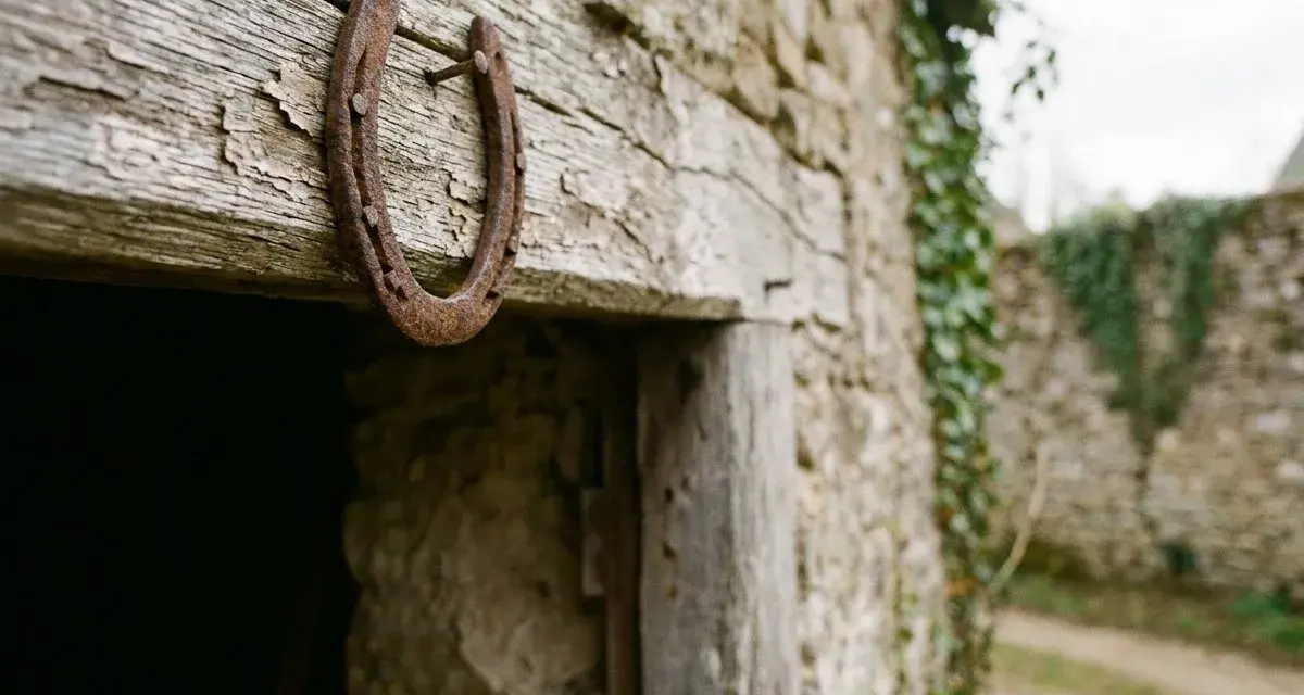 Un vieux fer à cheval pointes en bas, rouillé, est cloué sur une poutre en bois patiné, devant une bâtisse en pierre recouverte de lierre.