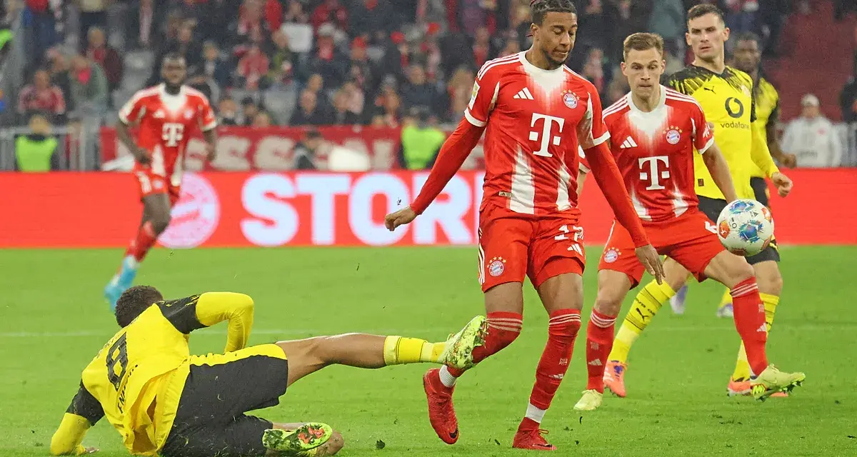 Intensywna akcja na boisku podczas meczu składy: Borussia Dortmund – Bayern Monachium. Piłkarz Bayernu w czerwonej koszulce próbuje odebrać piłkę leżącemu zawodnikowi BVB.