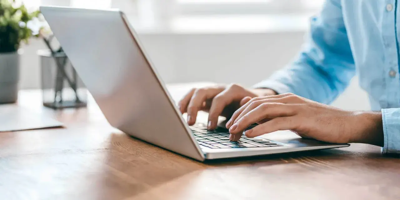 Person tippt auf einem silbernen Laptop auf einem Holztisch.