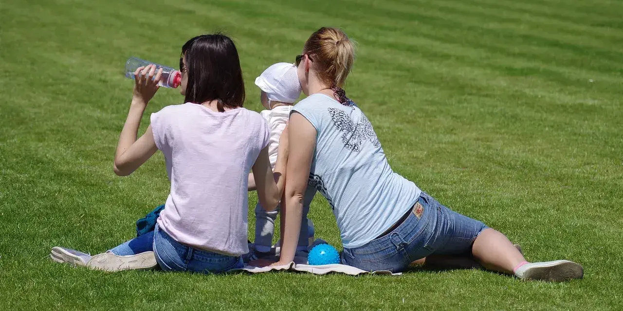 Dwie kobiety i dziecko siedzą na trawie. Jedna popija wodę, druga obejmuje dziecko. Widać brak zaufania po kłamstwie.