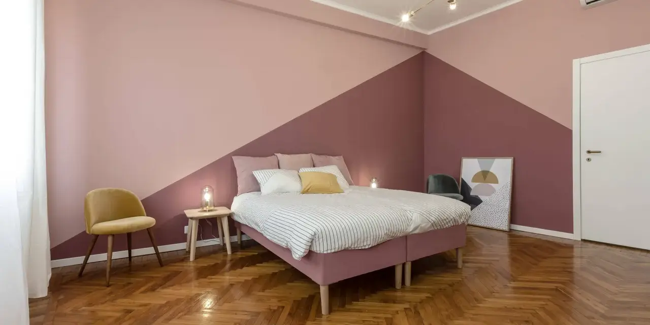 Camera da letto con pareti bicolore, un letto rosa, una sedia gialla e un parquet a spina di pesce. Un esempio di come pitturare una stanza con stile.