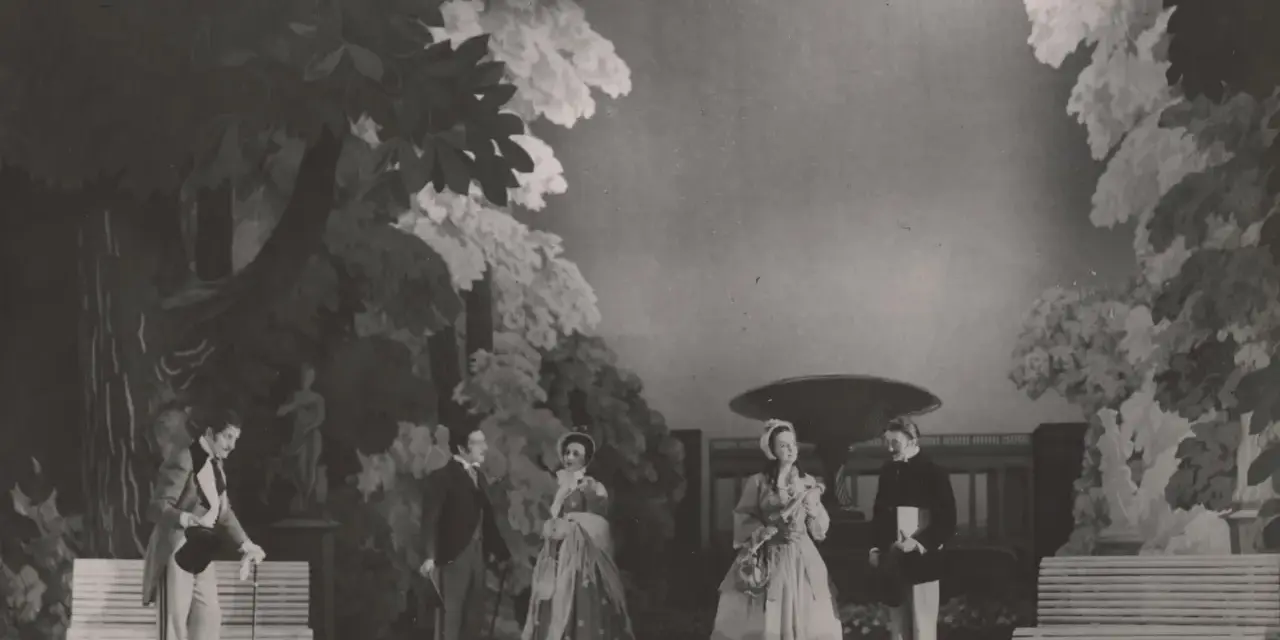 Scena z przedstawienia teatralnego, gdzie aktorzy w strojach z epoki odgrywają scenę w ogrodzie. Reżyser teatralny Szyfman zadbał o szczegóły scenografii.