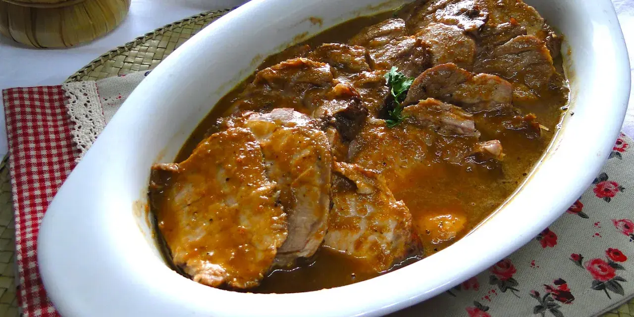 Arrosto di maiale morbido con sughetto, servito in una pirofila bianca, pronto per essere gustato.