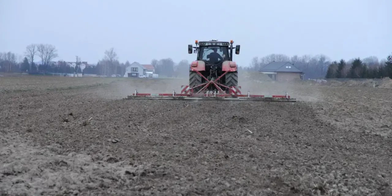 Czerwony traktor z agregatem uprawowym przygotowuje pole pod zasiew. Dzierżawa ziemi w pełni.