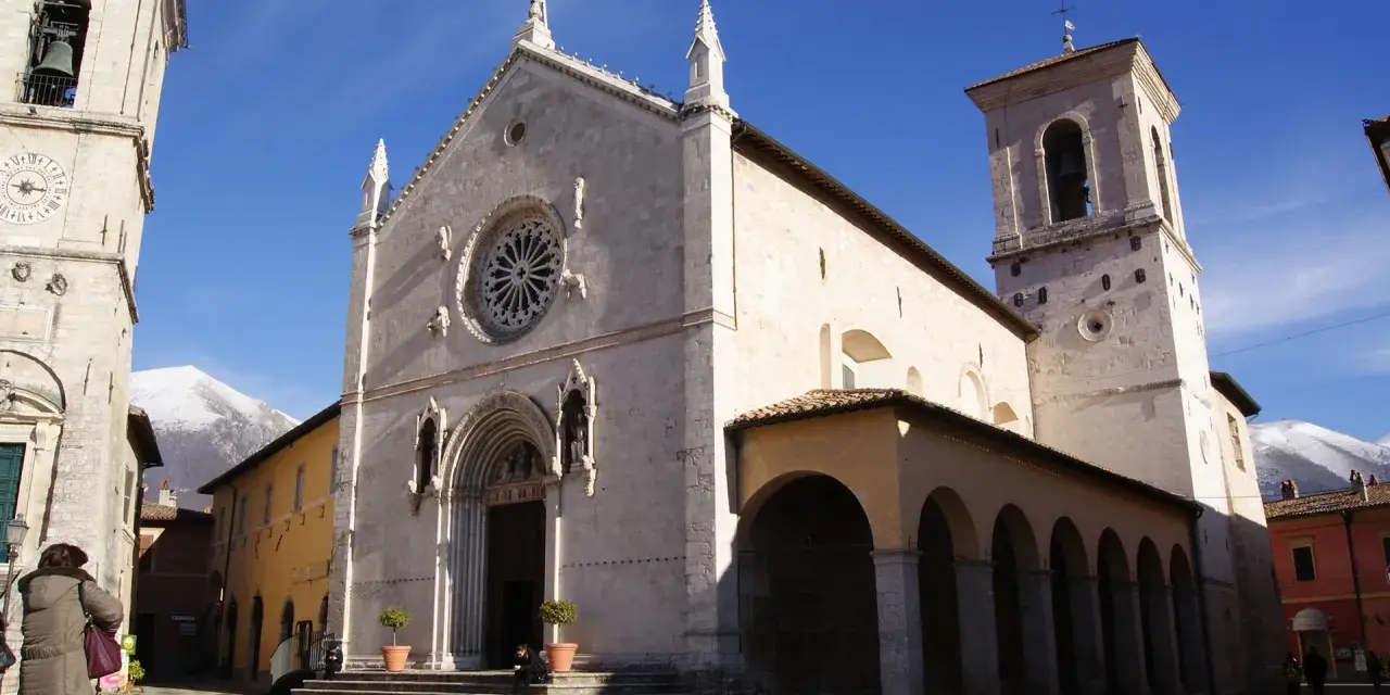 A grand church with a rose window and bell tower stands in a town square, with snow-capped mountains in the distance. This is Nursia, a historic Italian town.