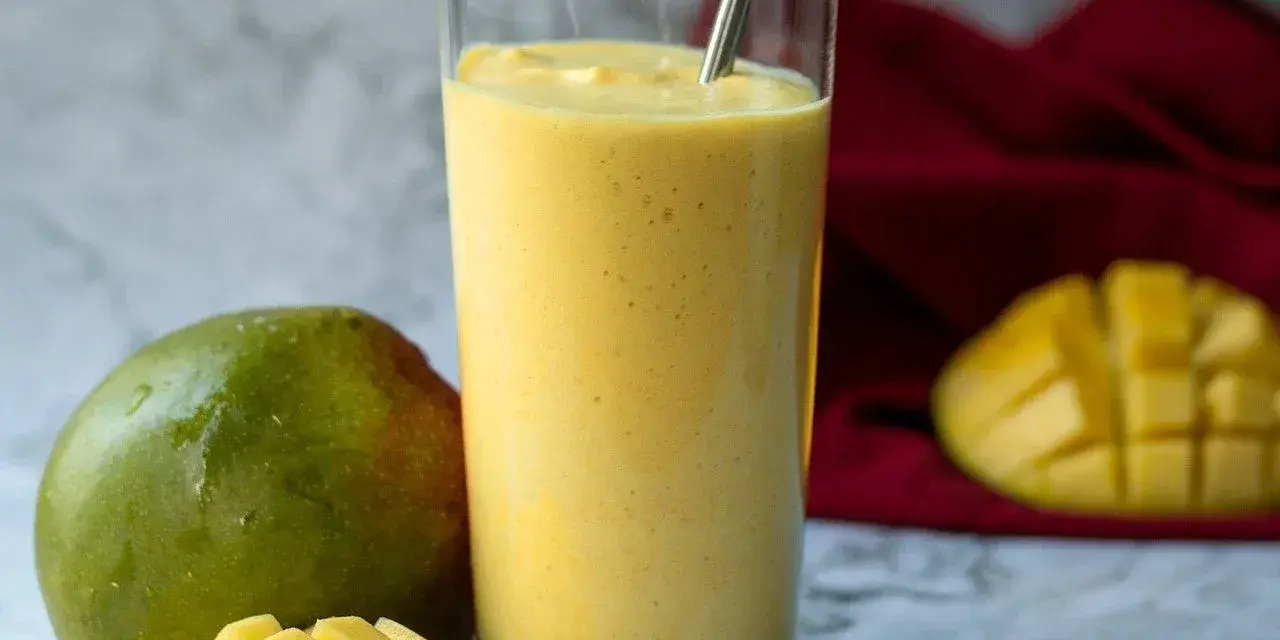 Orzeźwiający koktajl z mango w szklance, obok świeże mango i kawałki owocu. Idealny na upalne dni.