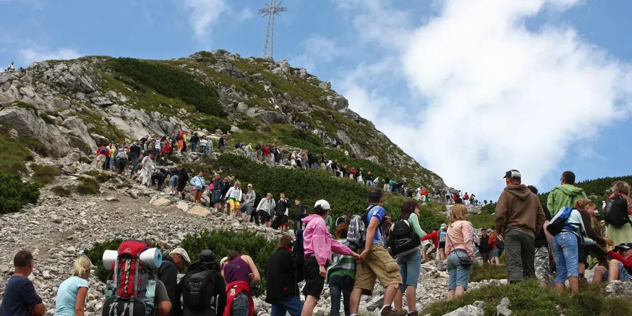 Tłum turystów wspina się na Giewont, podziwiając widok z Kasprowego Wierchu. Na szczycie widoczny krzyż.