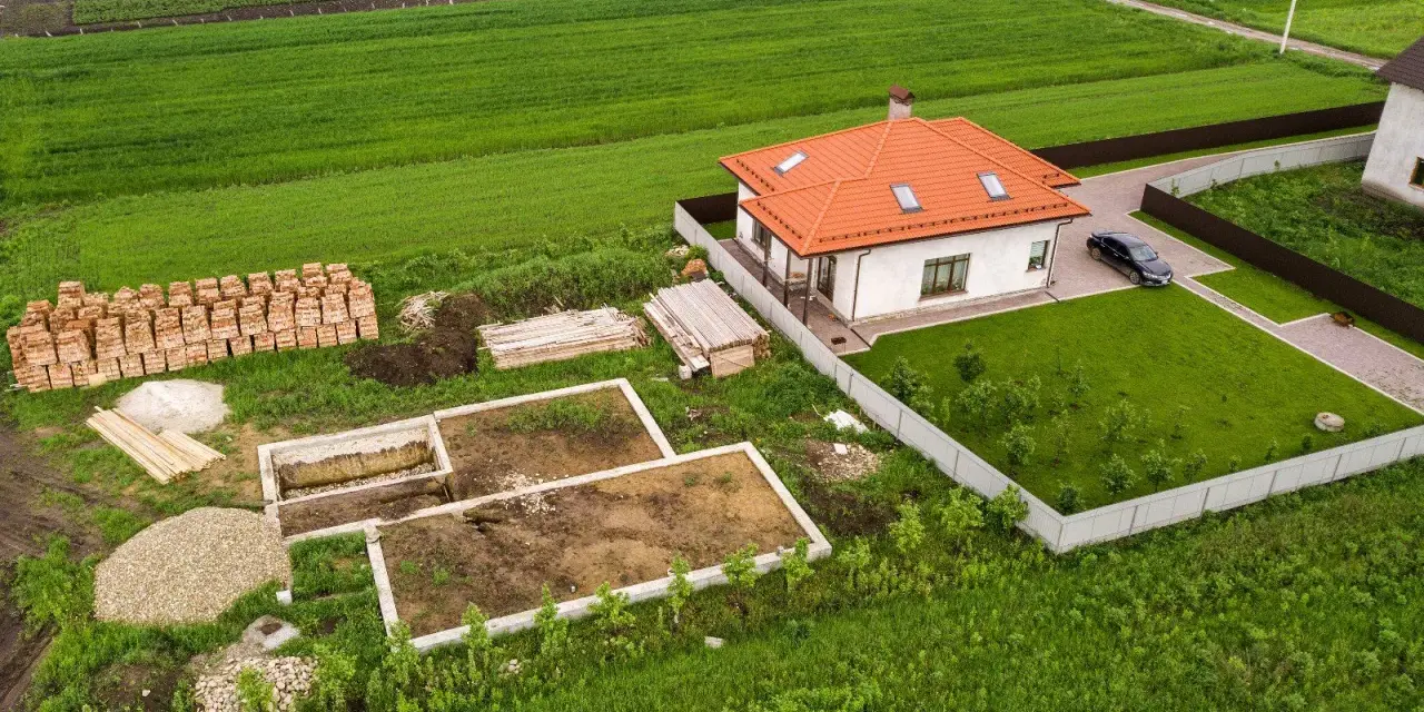 Działka 1000m2 wymiary: rozpoczęta budowa domu, gotowy fundament, sterta cegieł, drewno, żwir, obok stoi dom z pomarańczowym dachem i samochód.