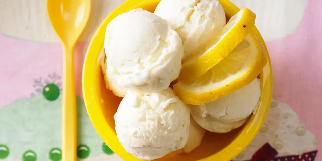 Erfrischendes Buttermilch Zitronen Eis in einer gelben Schale, garniert mit Zitronenscheiben. Ein gelber Löffel liegt daneben.