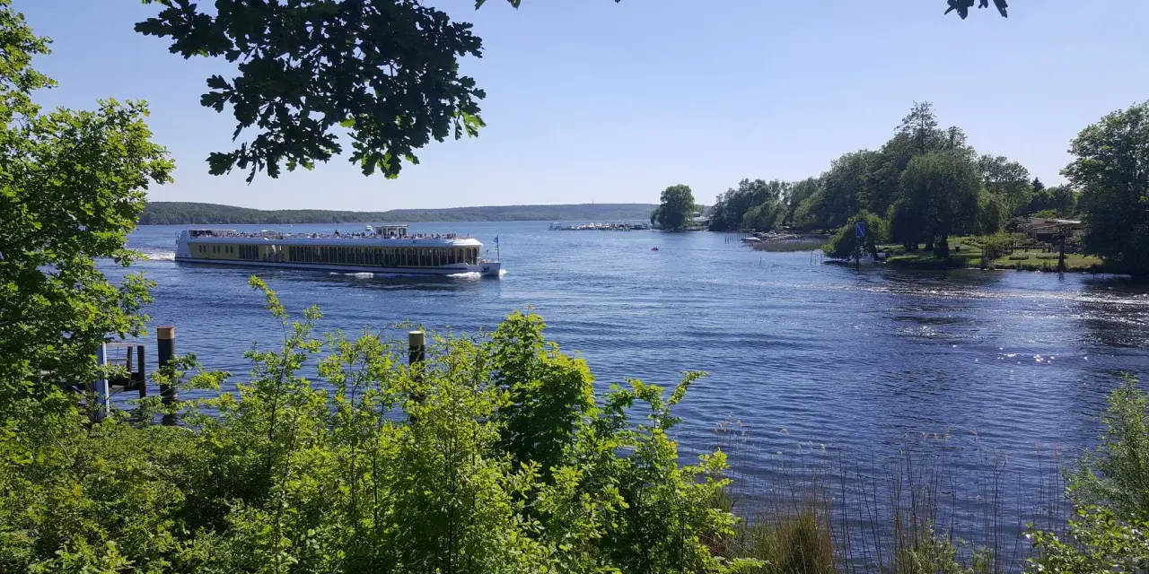 Ein Ausflugsboot fährt auf einem glitzernden See bei Potsdam. Grüne Bäume säumen das Ufer, ideal für einen entspannten Tag an den Badeseen Potsdam.