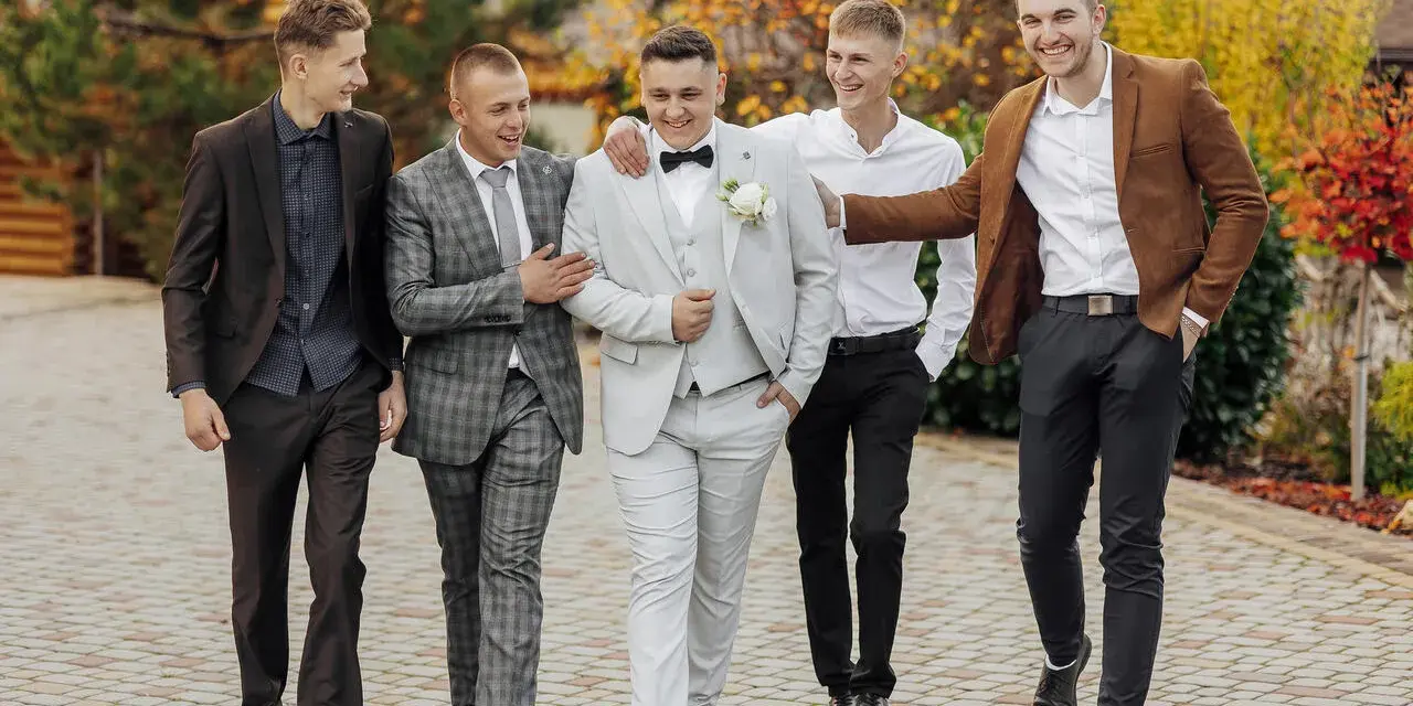 Pięciu mężczyzn w garniturach idzie razem. Jeden ma jasnoszary garnitur z satynowym połyskiem.