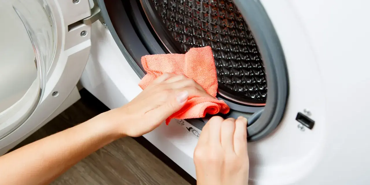 Hands cleaning the rubber seal of a washing machine with an orange cloth. This is part of how to clean washing machine.