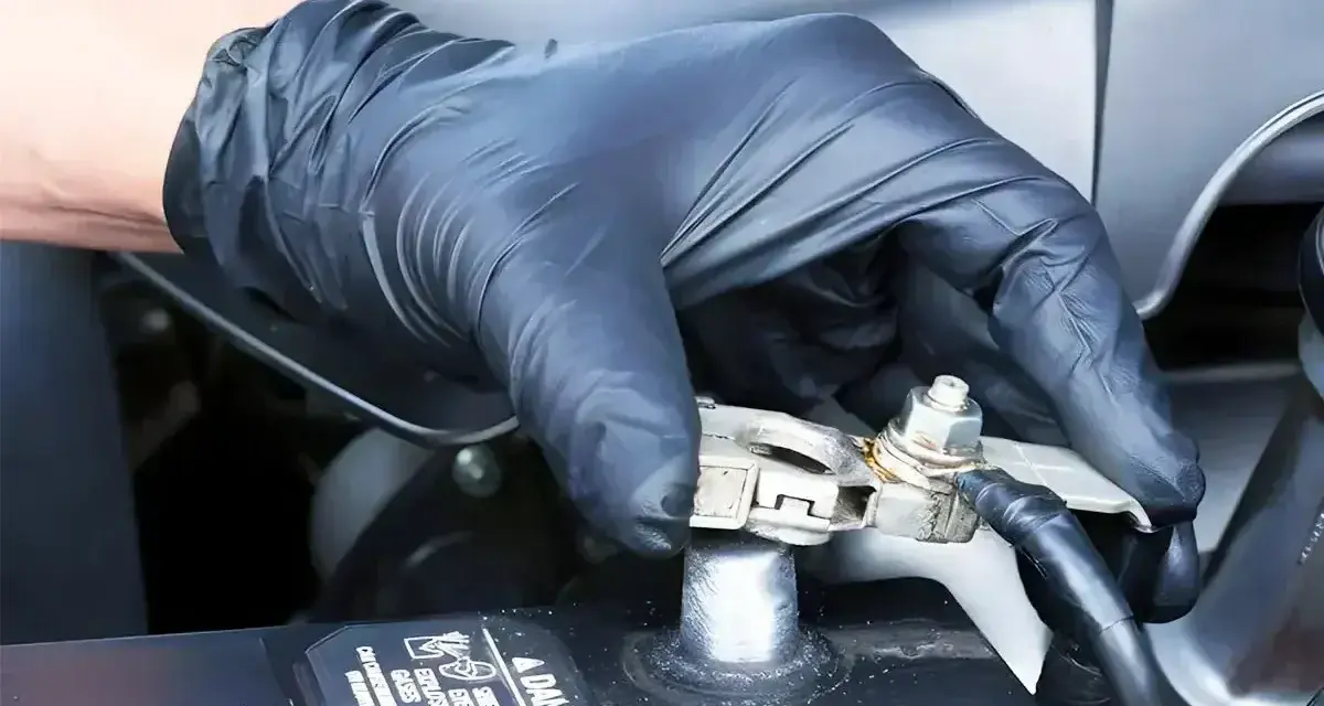 Gloved hand applying WD40 to a car battery terminal to prevent corrosion and ensure a good connection.