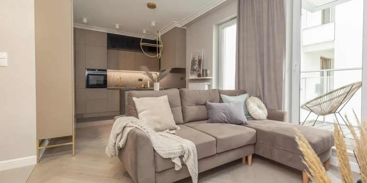 Elegancki salon z aneksem kuchennym w stylu monochromatycznym. Brązowa sofa z poduszkami, jasne drewno na podłodze.