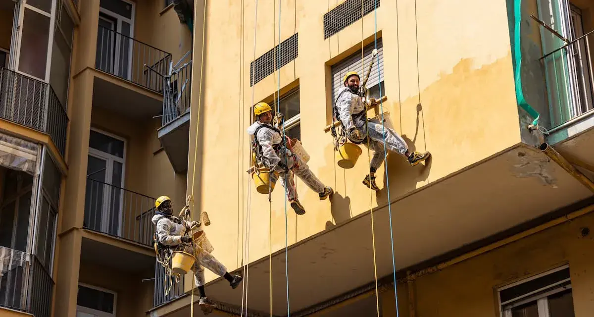Operai in corda applicano diversi tipi di intonaco per esterni su un edificio giallo.