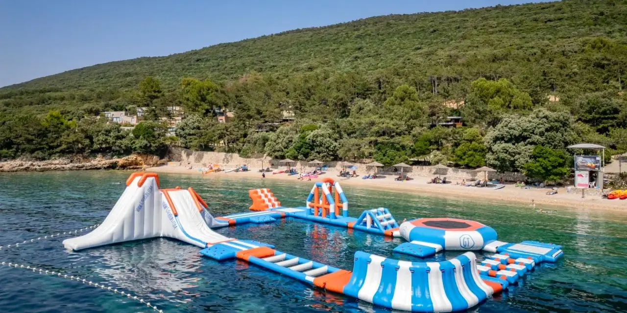 Wodne atrakcje na krystalicznie czystej wodzie, idealne na wakacje w **camping Slatina**. W tle zielone wzgórza i plaża.