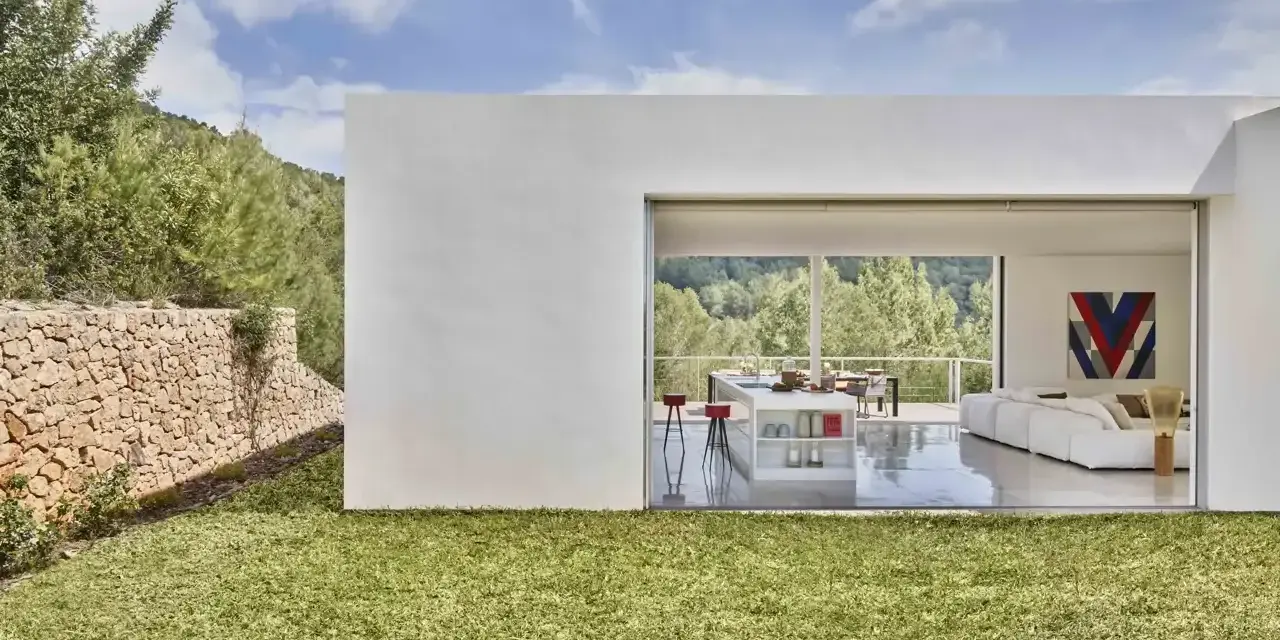 Exterior de casa ibicenca con césped verde y muro de piedra. Interior moderno con cocina y salón abiertos al paisaje.