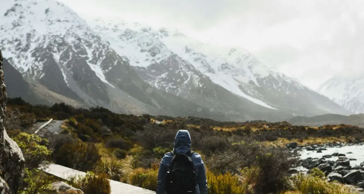 Une personne avec un sac à dos fait une randonnée sous la pluie, face à des montagnes enneigées. Le chemin en bois serpente à travers une végétation basse.
