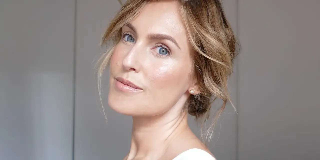 En kvinna med blont hår och blå ögon har ett naturligt glow i ansiktet. Hon bär en vit topp och en liten diamantörhänge.