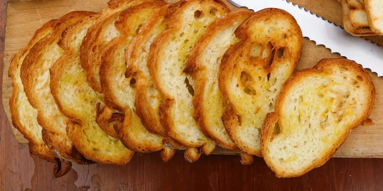 Fette dorate di pane con lievito madre, appena tostato, su un tagliere di legno. Un coltello seghettato è pronto per affettare altro pane.