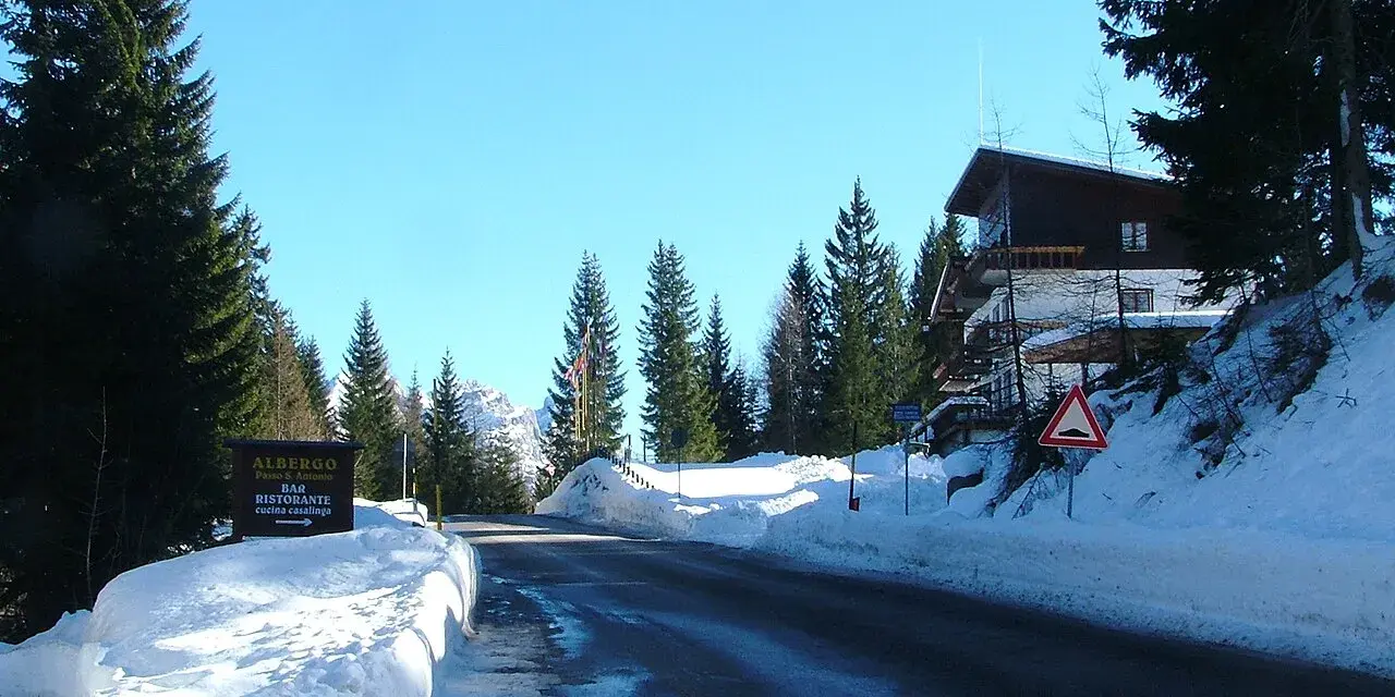 Strada innevata al passo San Antonio. Albergo e ristorante con cucina casalinga. Case di montagna e pini.