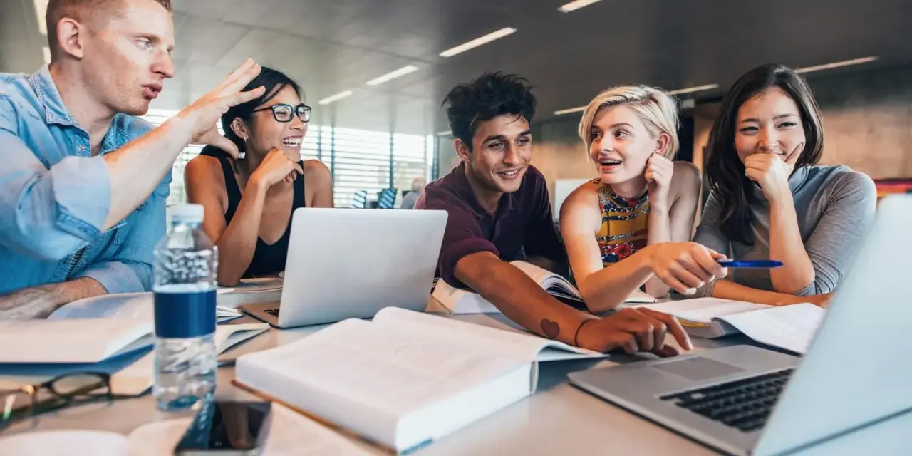 Fünf Studierende lernen gemeinsam. Sie lachen und diskutieren, während sie an Laptops arbeiten. Ein Mann gestikuliert lebhaft, was auf ein spannendes Studium im Ausland hindeutet.