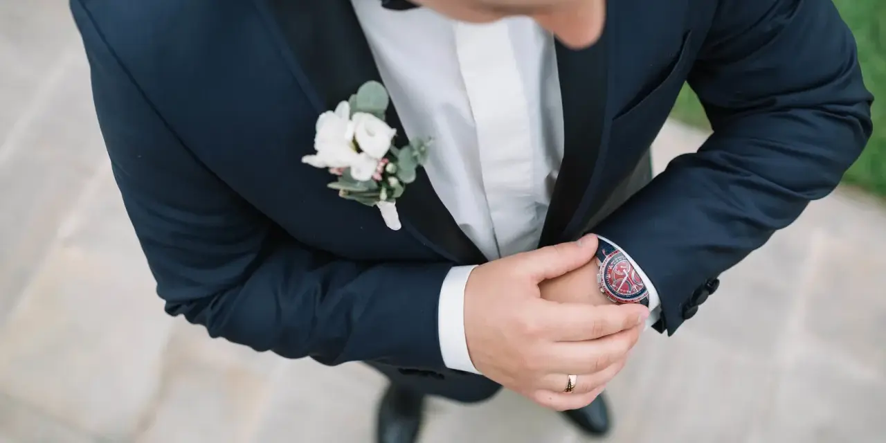 Elegancki pan młody w granatowym garniturze z butonierką co to jest, poprawia zegarek.