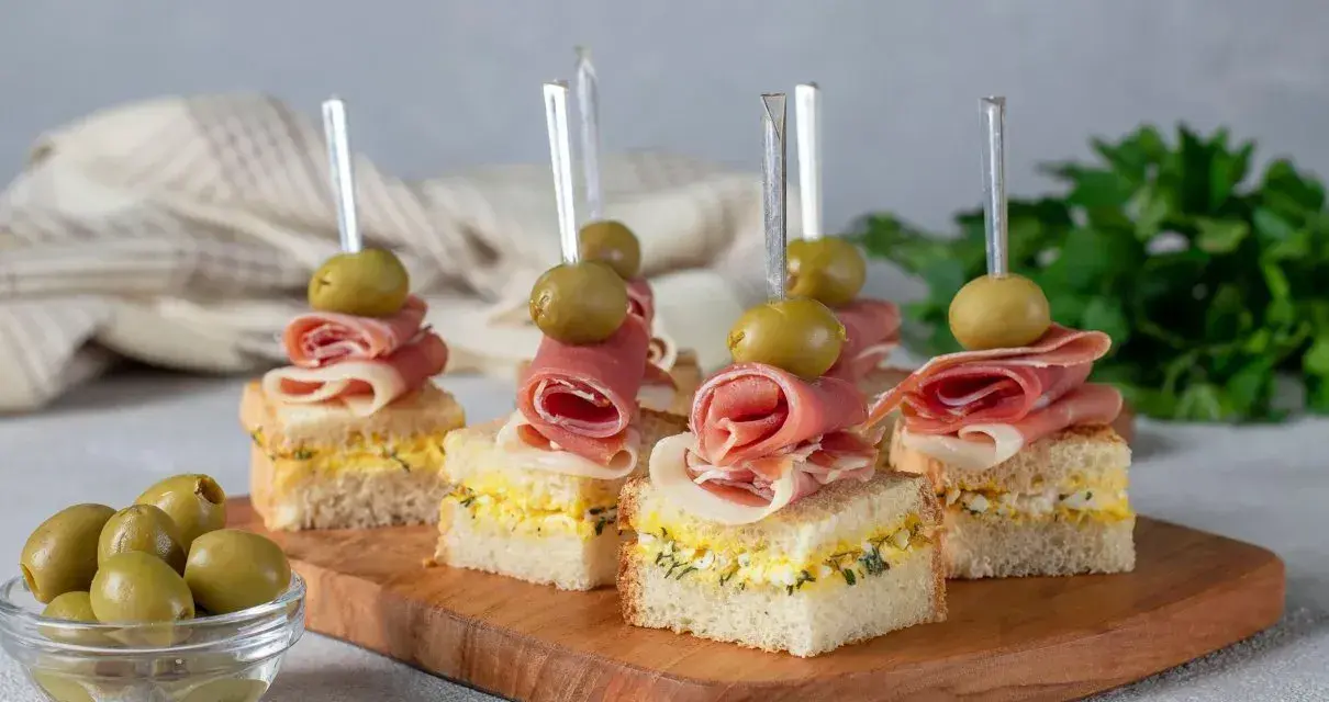Mini kanapki z szynką, jajkiem i oliwkami na drewnianej desce. Idealne finger food na przyjęcie.