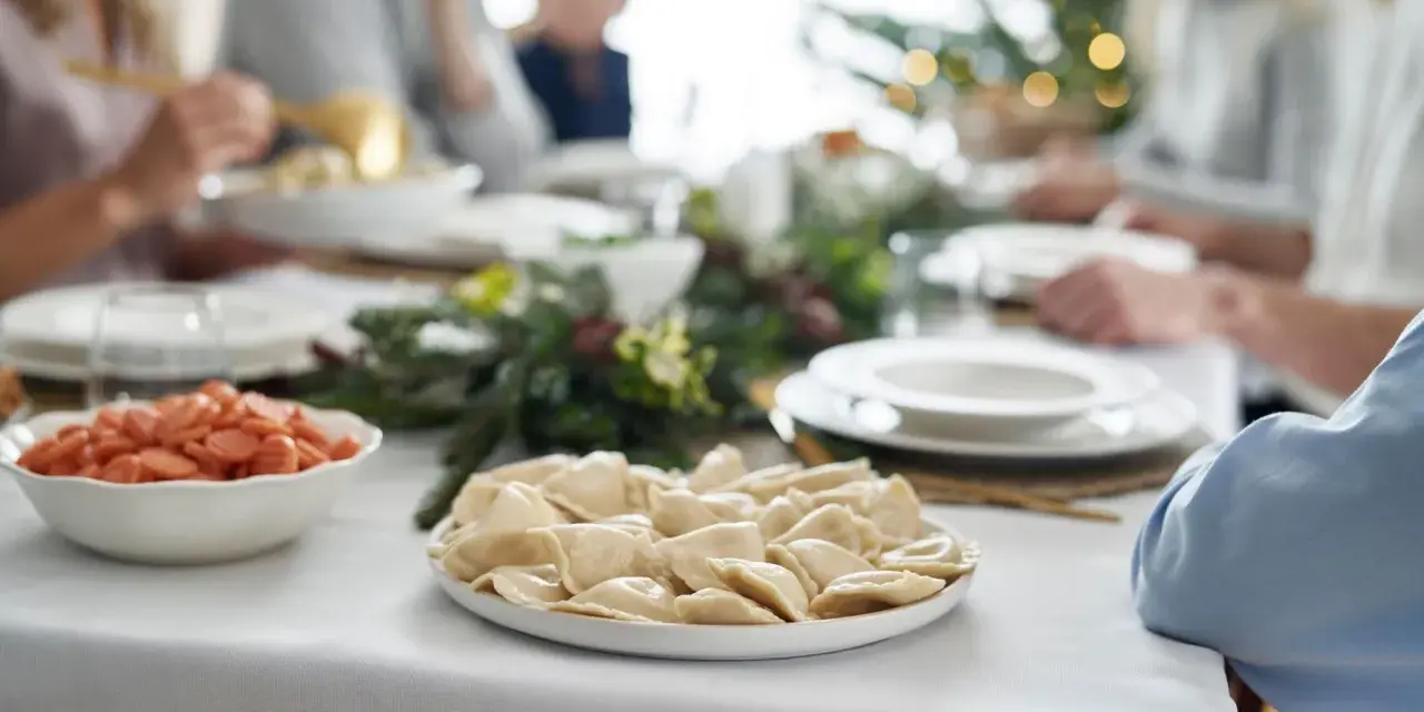 Talerz pierogów i miska z marchewką na wigilijnym stole, w tle rodzina i choinka.