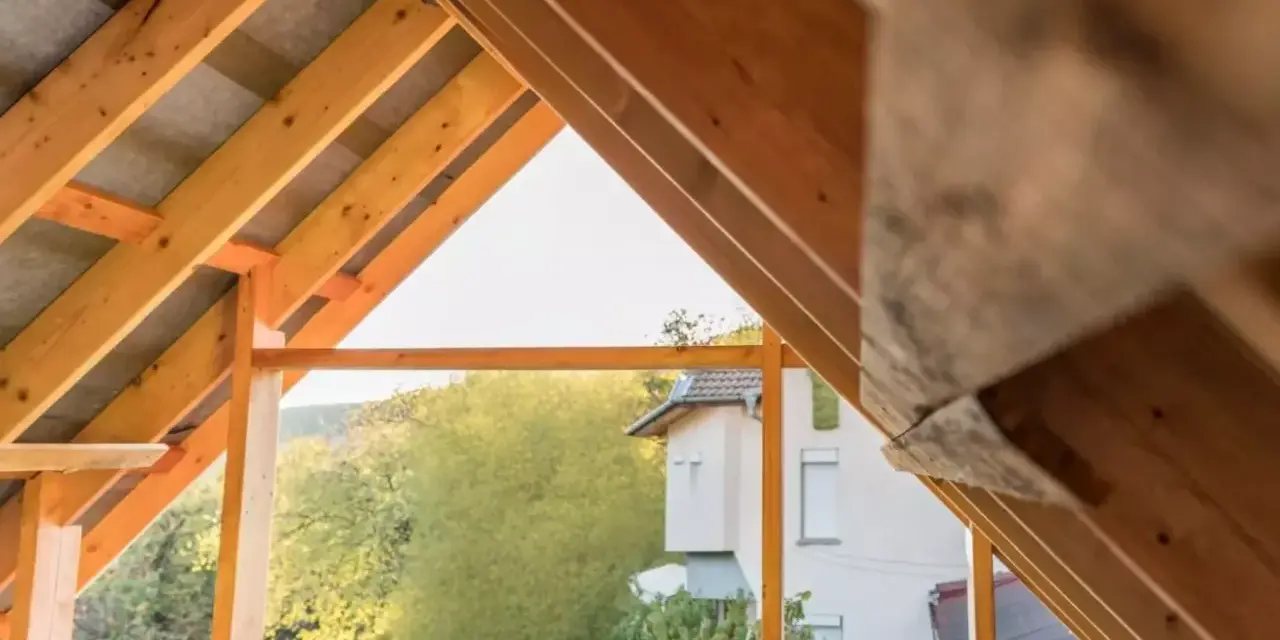 Drewniany szkielet dachu dwuspadowego w trakcie budowy. Widoczne krokwie i elementy konstrukcyjne, a w tle dom i zieleń.