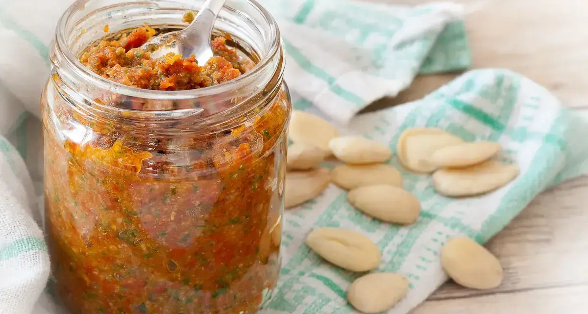 Vasetto di pesto di pomodori secchi, con mandorle sparse su un canovaccio.