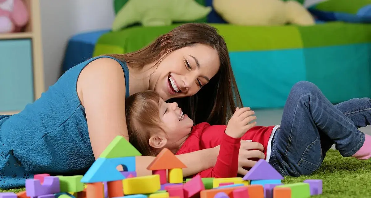 Uśmiechnięta opiekunka bawi się z dzieckiem wśród kolorowych klocków. Zastanawiasz się, ile kosztuje opieka nad dzieckiem za godzinę?