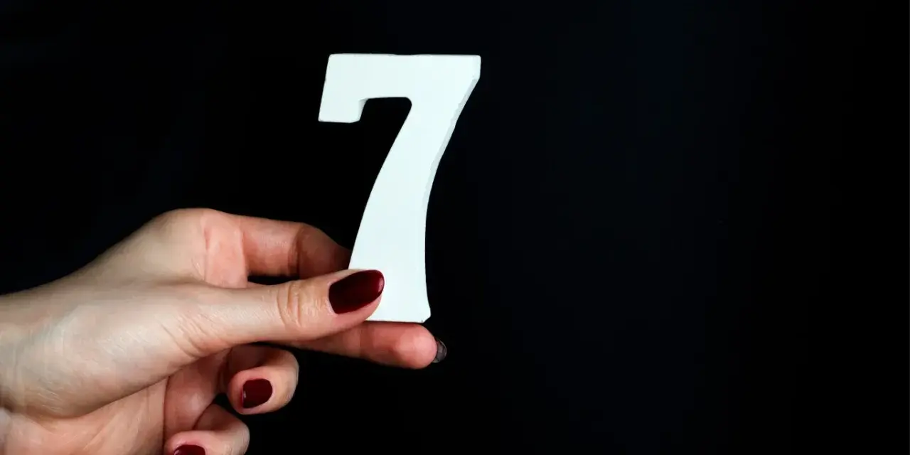 Dłoń z ciemnoczerwonym lakierem trzyma białą cyfrę 7. Może to symbolizować numerologiczną 7 mężczyzny, który jest tajemniczy i introspektywny.