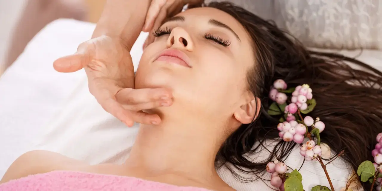 A woman receives an anti aging facial massage, her eyes closed in relaxation. Delicate pink berries adorn her dark hair.