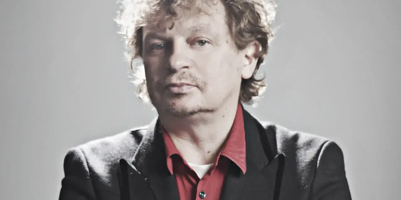 Marek Zieliński, aktor, w czarnej marynarce i czerwonej koszuli, z kręconymi włosami.