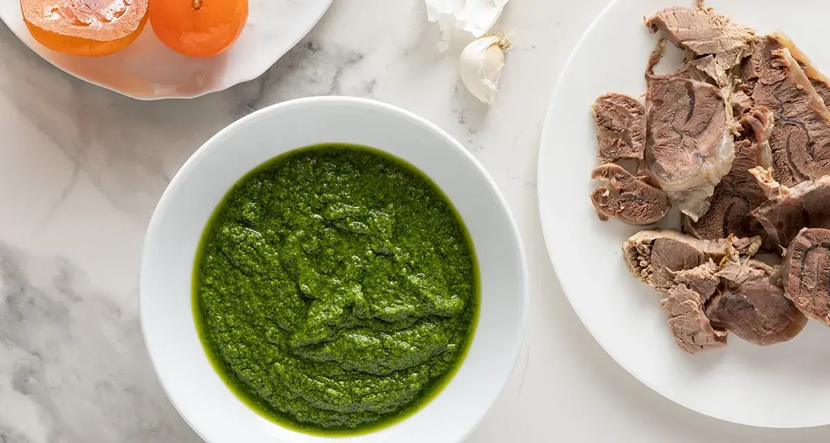 Una ciotola di vibrante salsa verde accanto a fette di carne bollita e pomodorini confit.