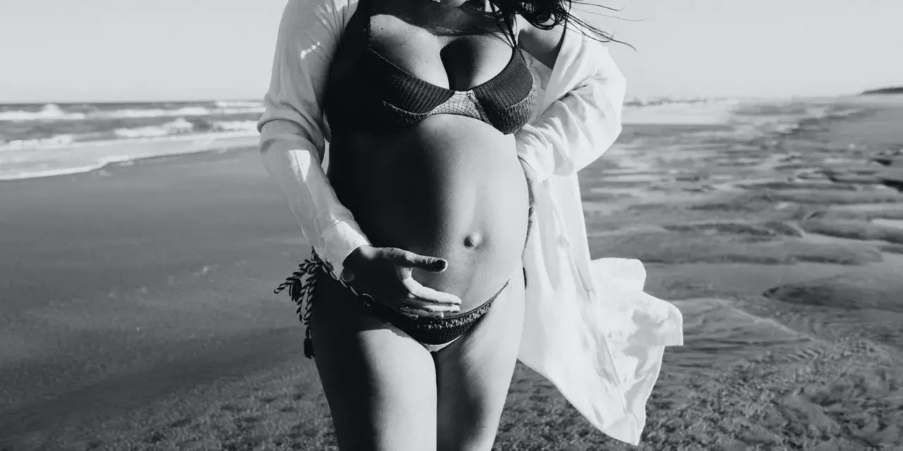 Donna incinta si gode il sole in spiaggia, accarezzando il pancione. La brezza marina le scompiglia i capelli.